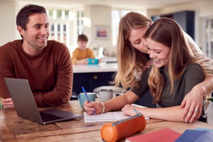 Parents Helping Teenage Daughter With Homework Sitting At Kitchen Table At Home Using Laptop