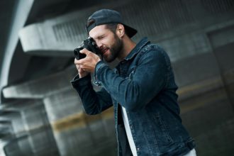 Photography hobby. Young stylish man standing on city street taking photos on camera concentrated